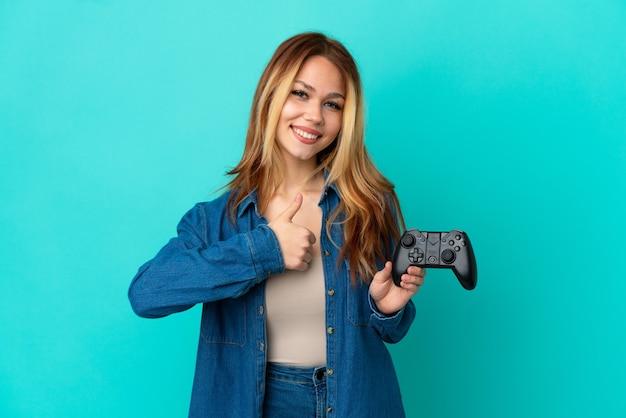 Tiener blond meisje speelt met een videogamecontroller over geïsoleerde muur en geeft een duim omhoog gebaar