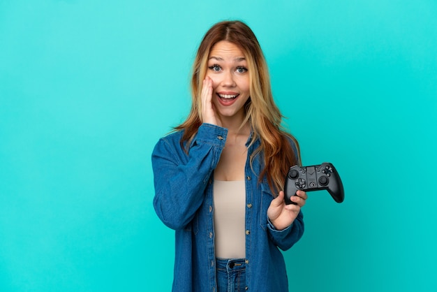 Tiener blond meisje speelt met een videogamecontroller over geïsoleerde muur met verrassing en geschokte gezichtsuitdrukking