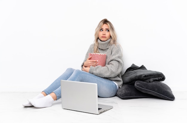 Tiener blondevrouw die popcorn eten terwijl het letten van op een film op laptop die twijfelsgebaar maken terwijl het opheffen van de schouders