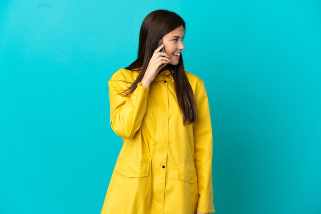 Tiener Braziliaans meisje dat een regendichte laag over geïsoleerde blauwe achtergrond draagt die een gesprek met de mobiele telefoon met iemand houdt