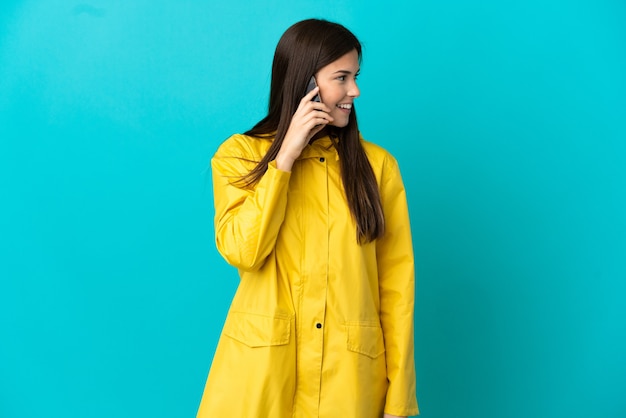 Tiener braziliaans meisje dat een regendichte laag over geïsoleerde blauwe achtergrond draagt die een gesprek met de mobiele telefoon met iemand houdt