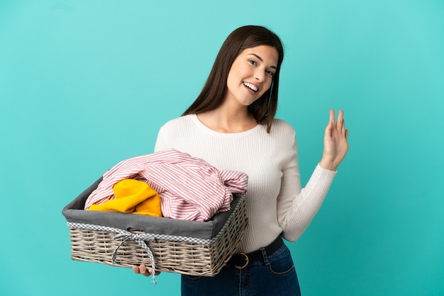 Tiener Braziliaans meisje met een wasmand geïsoleerd op blauwe achtergrond saluerend met de hand met gelukkige uitdrukking