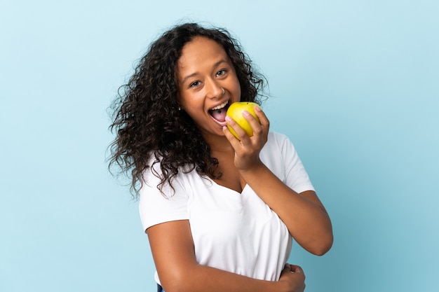 Tiener Cubaans meisje dat op blauwe muur wordt geïsoleerd die een appel eet