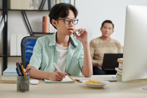 Tiener die chips eet en online lessen op de computer bekijkt terwijl hij thuis studeert, zijn vader werkt op de achtergrond