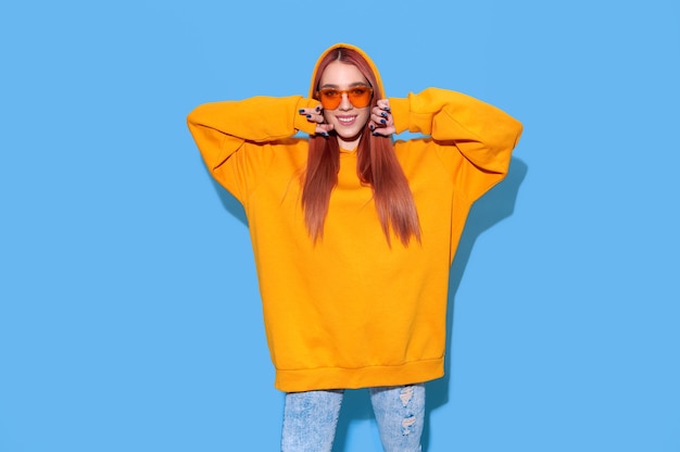 Tiener hipster meisje in oranje oversized hoodie en trendy zonnebril op heldere blauwe achtergrond