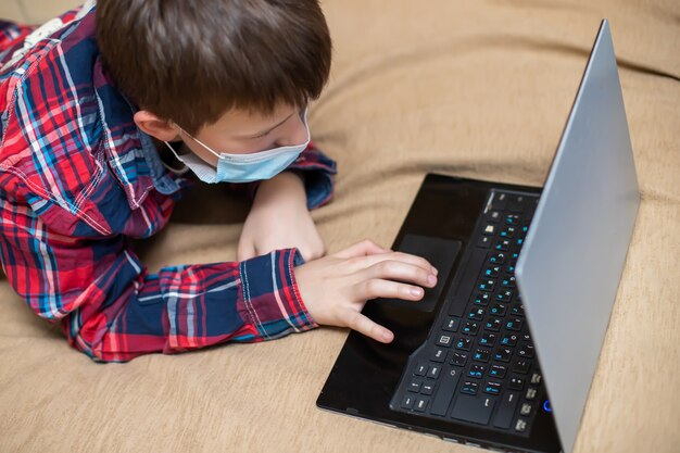 Tiener in beschermende medische masker hoest in vuist. kind doet op afstand lessen liggend op bed in de buurt van laptop. concept van het leren en spelen van kinderen op de computer.