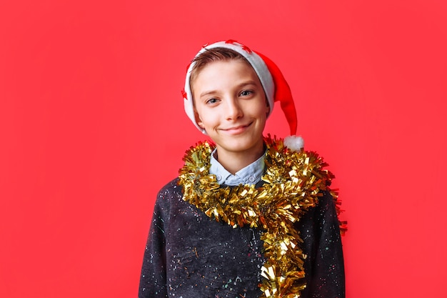 Tiener in Kerstmanhoed en met klatergoud op hals die op rode muur glimlachen