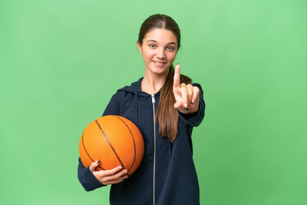 Tiener kaukasisch meisje dat basketbal over geïsoleerde achtergrond speelt die een vinger toont en opheft