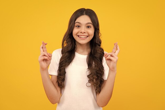 Tiener kind met vingers gekruist voor goed geluk portret van vrolijk meisje bidt en hoopt dat dromen uitkomen kruist vingers voor goed geluk geïsoleerd op gele studio achtergrond