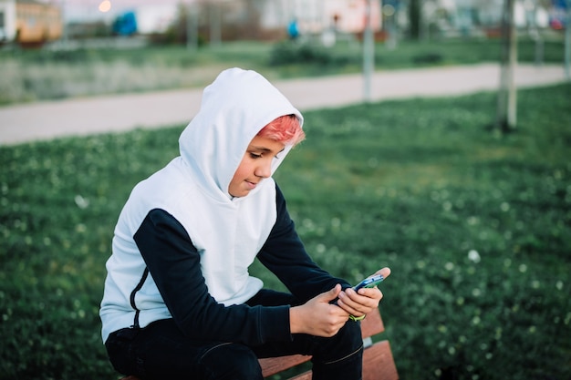 Tiener met een kap die mobiel in een park bekijkt