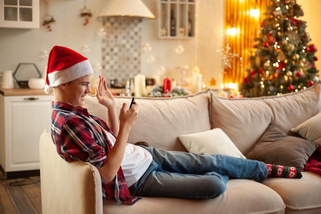 Tiener met kerstmuts zit in een met kerst versierde kamer en chat online met een jongen met een smartphone