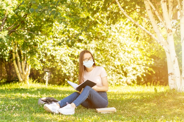 Tiener met maskerstudent op het gras in het park met boeken en een rugzak terug naar school na het coronavirus