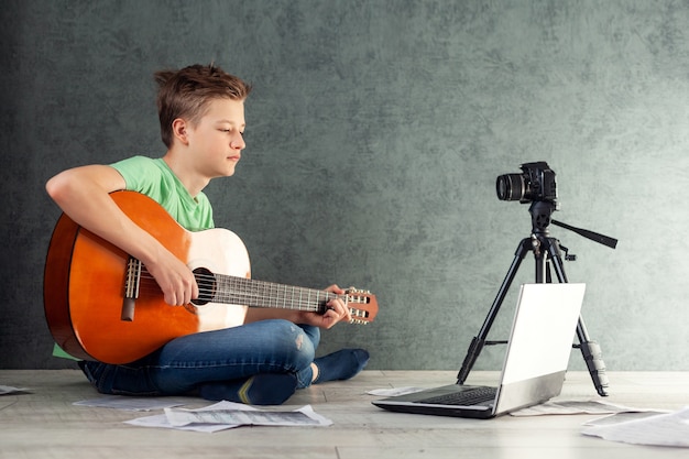 Tiener neemt de vlog van een spel op een gitaar thuis op de camera af. Jonge videobloger tienerjongen leert gitaar spelen online in de woonkamer,