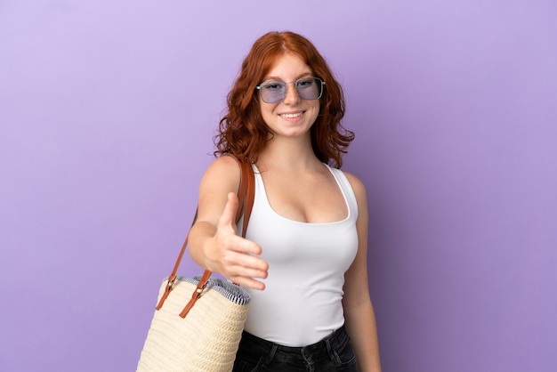 Tiener roodharig meisje met een strandtas geïsoleerd op paarse achtergrond handen schudden voor het sluiten van een goede deal