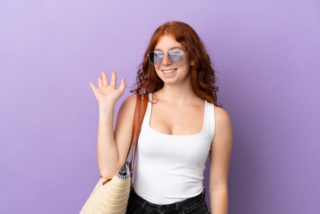 Tiener roodharig meisje met een strandtas geïsoleerd op paarse achtergrond saluerend met de hand met gelukkige uitdrukking