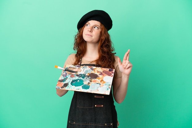 Tiener roodharige artiest met een palet geïsoleerd op een groene achtergrond met vingers die elkaar kruisen en het beste wensen