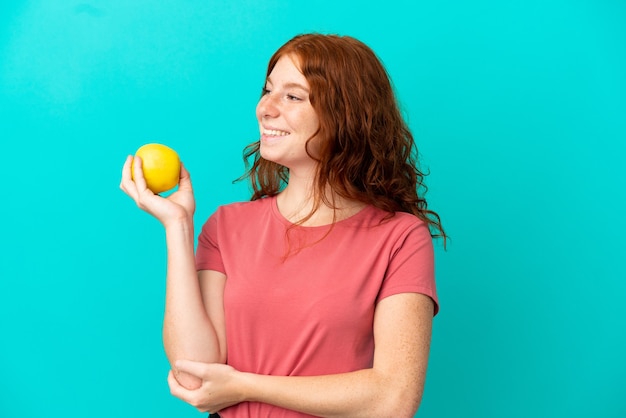 Tiener roodharige meisje geïsoleerd op blauwe achtergrond met een appel en happy