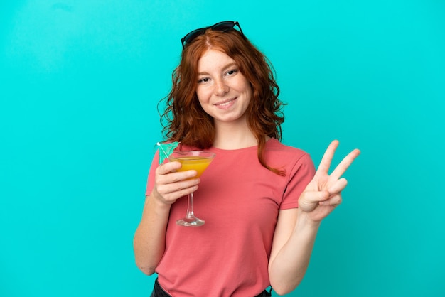 Tiener roodharige meisje met cocktail geïsoleerd op blauwe achtergrond glimlachend en overwinning teken tonen
