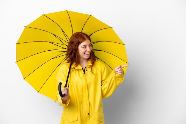 Tiener roodharige meisje regendichte jas en paraplu geïsoleerd op een witte achtergrond wijzende vinger naar de zijkant en de presentatie van een product