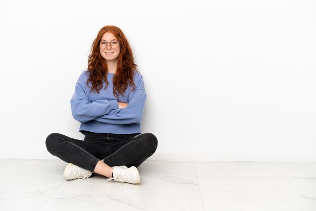 Foto tiener roodharige meisje zittend op de vloer geïsoleerd op een witte achtergrond met de armen gekruist in frontale positie