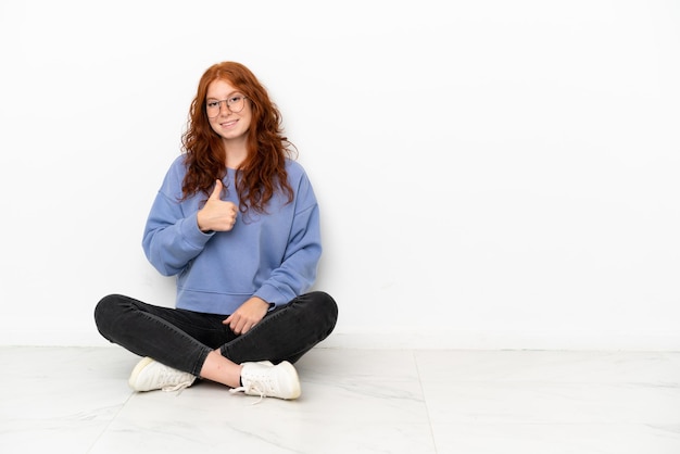 Tiener roodharige meisje zittend op de vloer geïsoleerd op een witte achtergrond met een duim omhoog gebaar