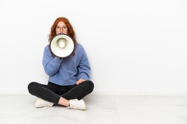 Tiener roodharige meisje zittend op de vloer geïsoleerd op een witte achtergrond schreeuwen door een megafoon