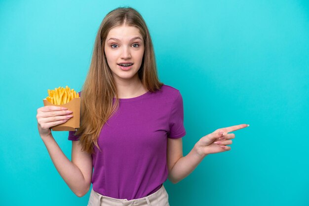 Tiener Russisch meisje dat frietjes vangt die op een blauwe achtergrond worden geïsoleerd, verrast en met de vinger naar de zijkant wijst