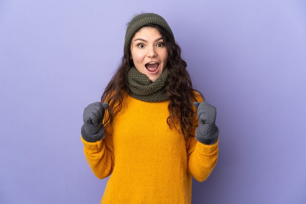 Tiener Russisch meisje dat met de winterhoed op purpere muur wordt geïsoleerd die een overwinning in winnaarspositie viert