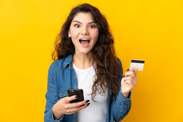 Tiener Russisch meisje dat op gele muur wordt geïsoleerd die met mobiel koopt en een creditcard met verbaasde uitdrukking houdt
