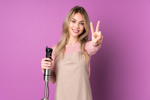 Tiener Russisch meisje die handmixer op purpere muur gebruiken die en overwinningsteken glimlachen tonen