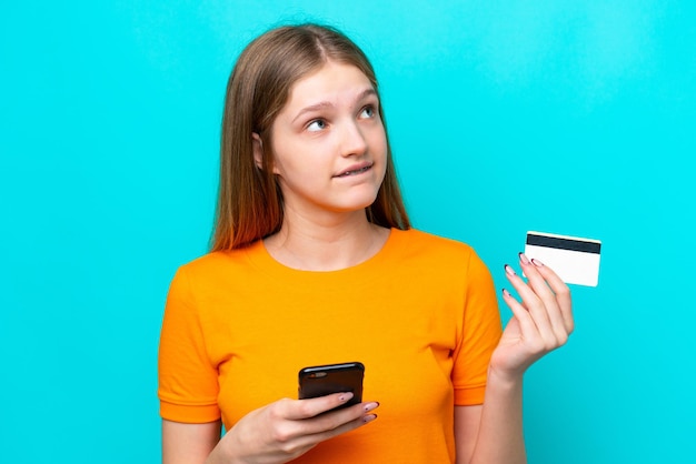 Tiener Russisch meisje geïsoleerd op blauwe achtergrond kopen met de mobiel met een creditcard tijdens het denken