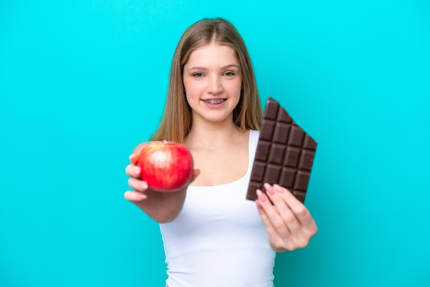 Tiener Russisch meisje geïsoleerd op een blauwe achtergrond die een chocoladetablet in de ene hand en een appel in de andere neemt