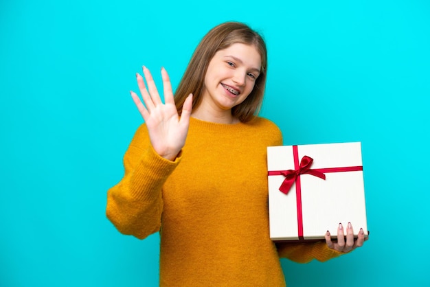 Tiener Russisch meisje met een geschenk geïsoleerd op blauwe achtergrond saluerend met de hand met gelukkige uitdrukking