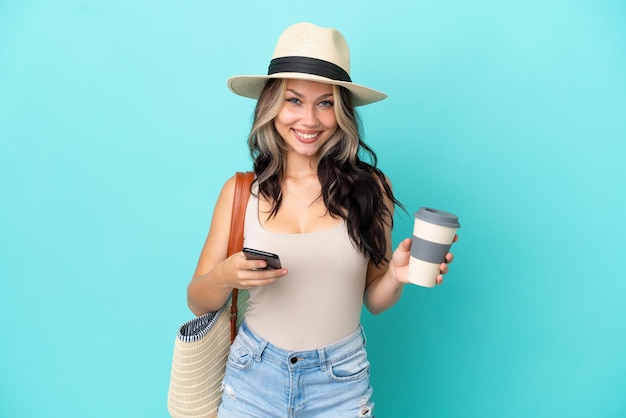 Tiener Russisch meisje met pamel en strandtas geïsoleerd op blauwe achtergrond met koffie om mee te nemen en een mobiel