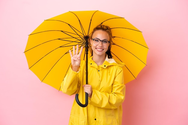 Tiener Russisch meisje met regenbestendige jas en paraplu geïsoleerd op roze achtergrond gelukkig en vier tellen met vingers
