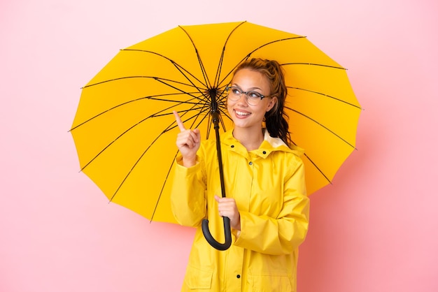 Tiener Russisch meisje met regenbestendige jas en paraplu geïsoleerd op roze achtergrond met de bedoeling de oplossing te realiseren terwijl ze een vinger optilt