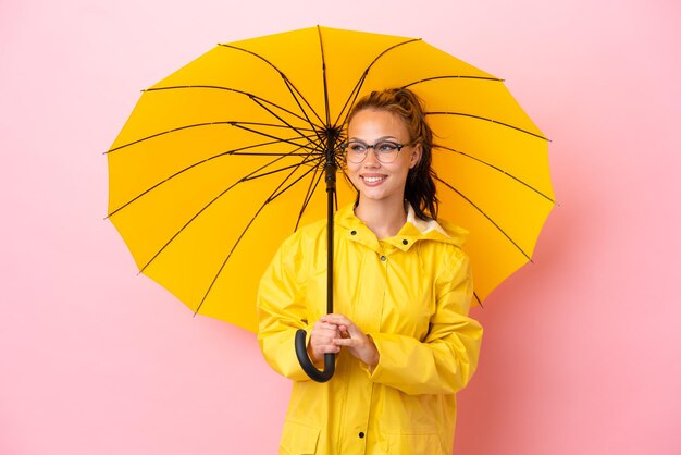 Tiener Russisch meisje met regenbestendige jas en paraplu geïsoleerd op roze achtergrond op zoek naar de zijkant en glimlachen