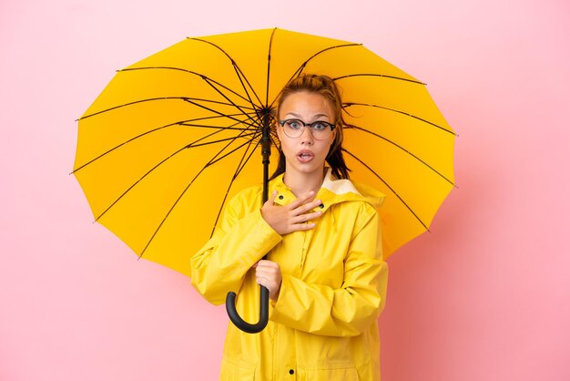 Tiener Russisch meisje met regenbestendige jas en paraplu geïsoleerd op roze achtergrond verrast en geschokt terwijl ze naar rechts kijkt