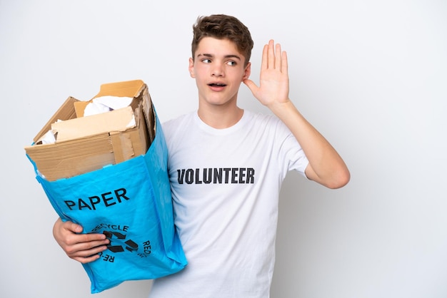 Tiener russische man met een recyclingzak vol papier om te recyclen geïsoleerd op een witte achtergrond, luisterend naar iets door hand op het oor te leggen