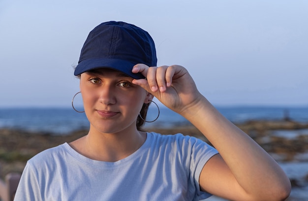 Tiener staat bij de zee bij zonsondergang en kijkt recht naar de camera. tienermeisje dat een t-shirt en een donkerblauwe baseballpet draagt en het vizier aanraakt. mockup voor pet en t-shirt