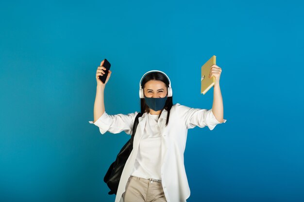 Tiener vrouw die met rugzak gezichtsmasker en hoofdtelefoons draagt die gele blocnote en telefoon op blauw houden