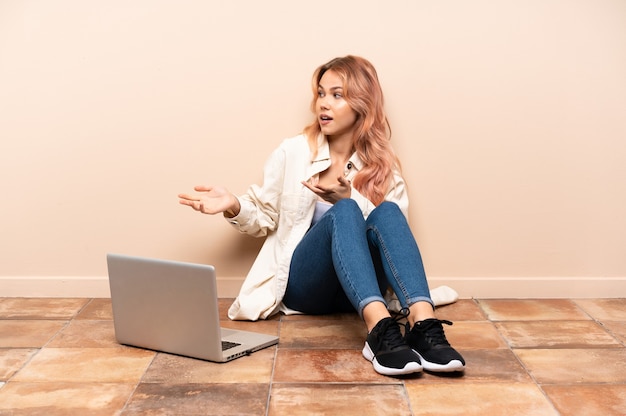 Tiener vrouw met een laptop zittend op de vloer binnenshuis met verrassing expressie terwijl op zoek naar kant