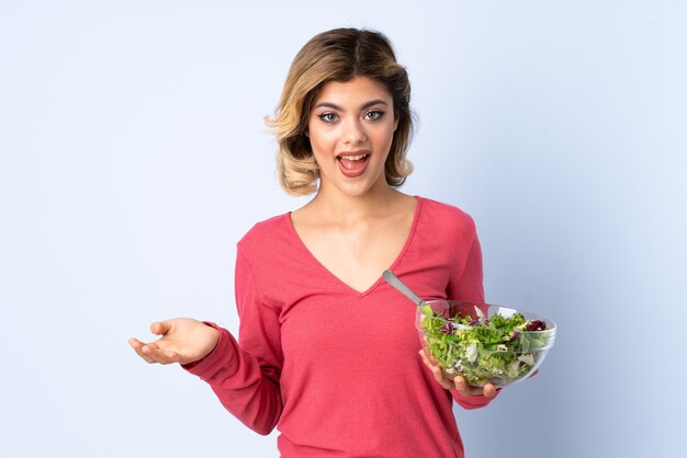 Tiener vrouw met salade geïsoleerd op blauw met geschokte gelaatsuitdrukking