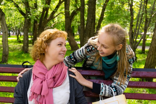 Tienerdochter loopt achter haar moeder aan zittend op een bankje in het park en knuffelt plotseling haar schouders