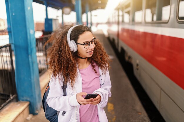 Tienerhipstermeisje met hoofdtelefoons en slim telefoon in hand wachten om op trein binnen te komen.