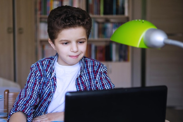 Tienerjongen aan een tafel met een laptop kijkt vreugdevol naar de monitor Coronavirus in quarantaine geplaatst thuisonderwijs Selectieve focus Achtergrond vervagen