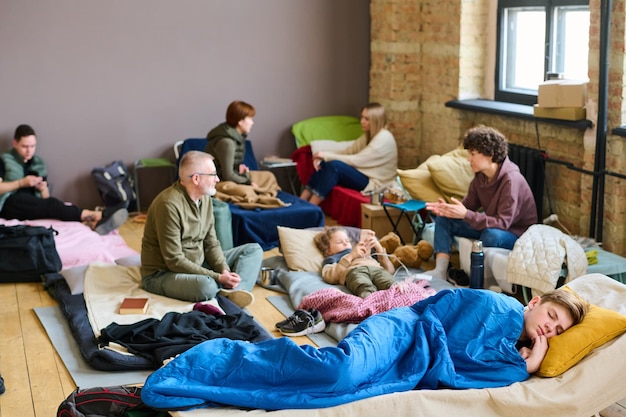 Tienerjongen die op de bank slaapt tegen andere vluchtelingen die op bedden praten