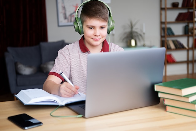 Tienerjongen in hoofdtelefoons die online studeren met behulp van laptop die in notitieboekje schrijft.