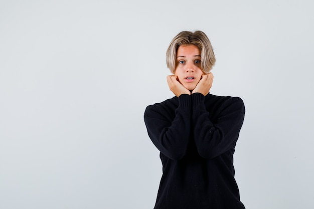 Tienerjongen in zwarte sweater die kin op handen leunt en verrast kijkt, vooraanzicht.