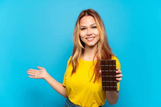 Tienermeisje dat chocola vasthoudt over een geïsoleerde blauwe achtergrond die de handen naar de zijkant uitstrekt om uit te nodigen om te komen